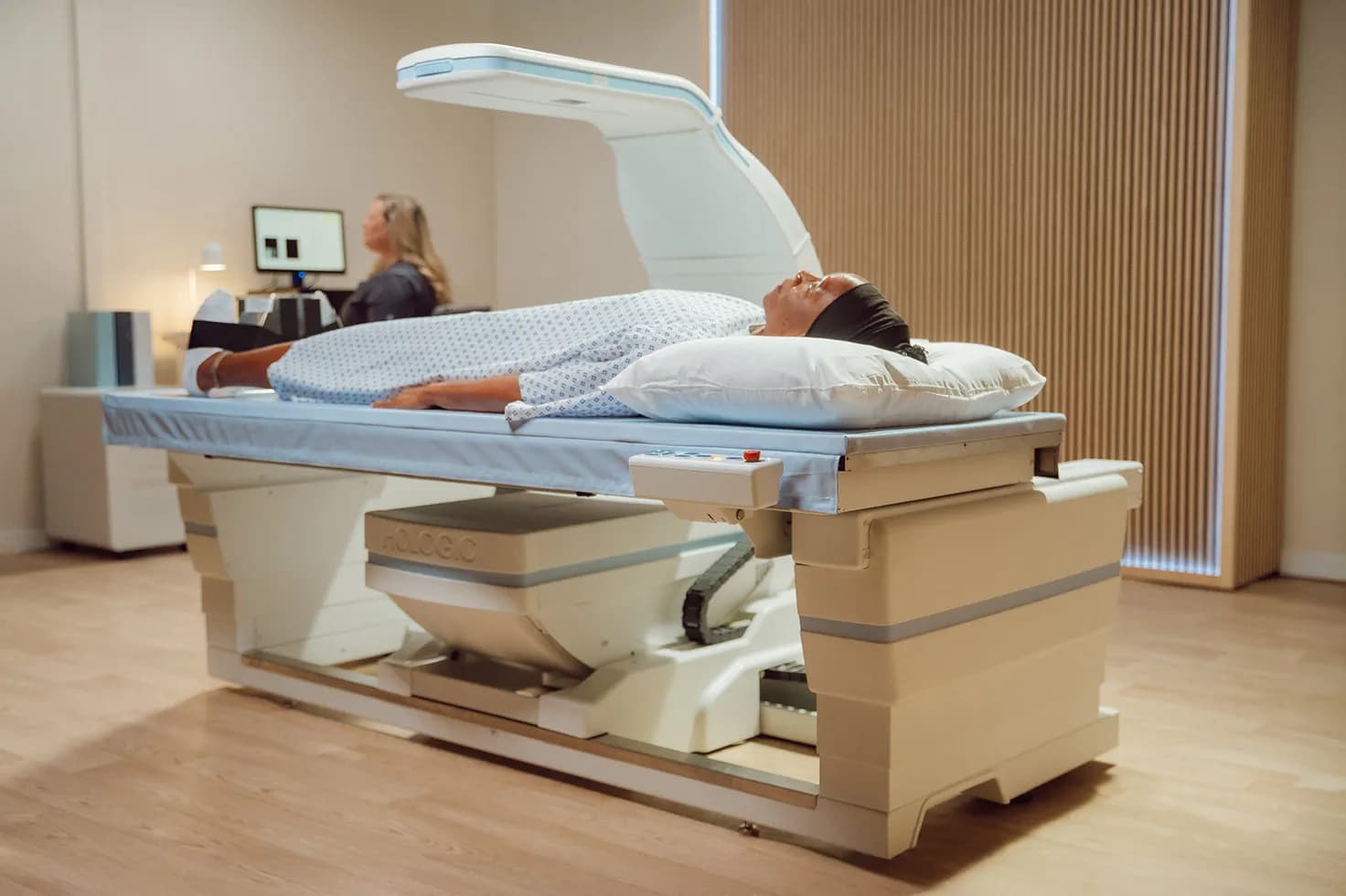 A person lying on the open, padded table of a DEXA scanner, with the scanning arm suspended above the torso and a technologist seated at the control console behind.